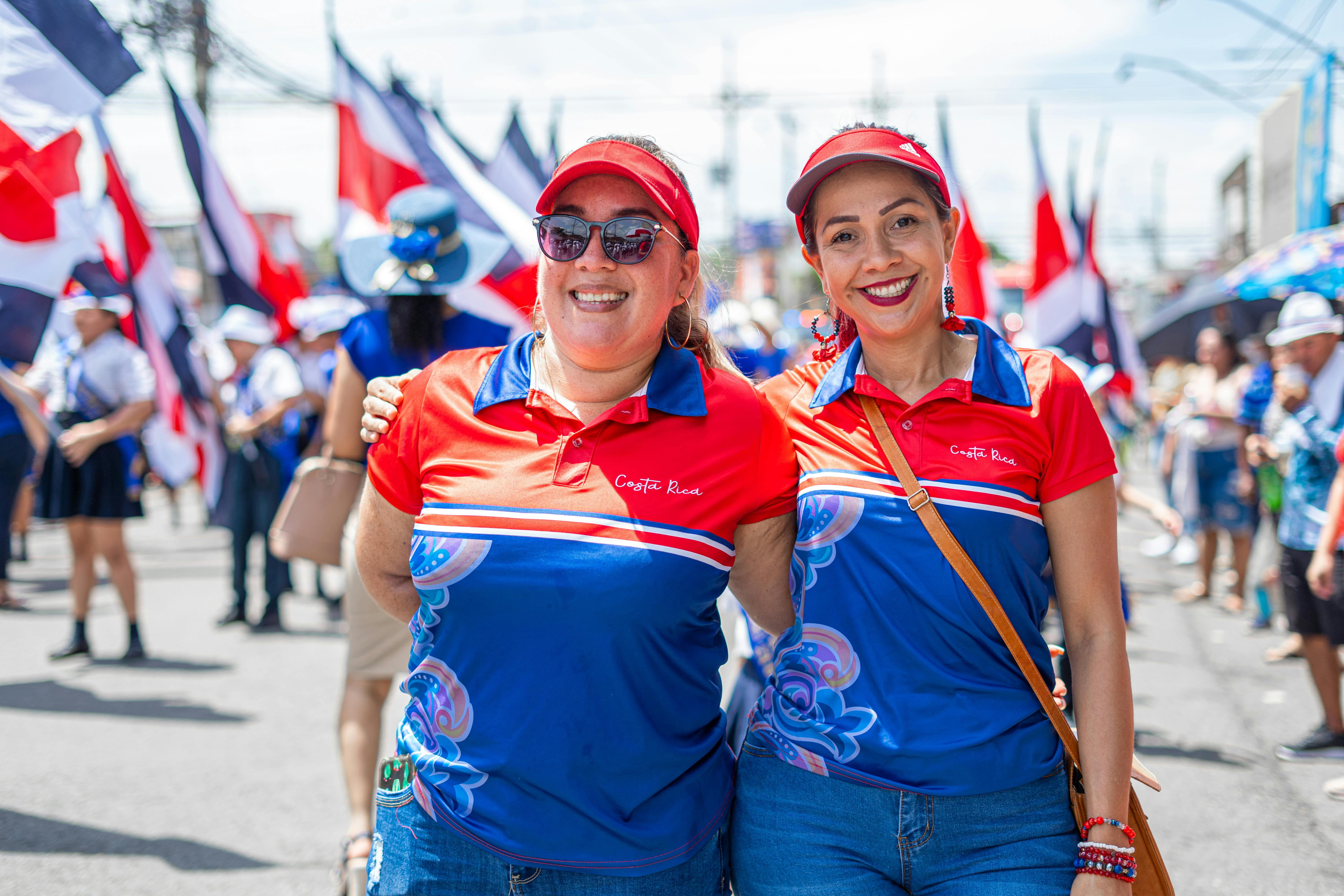 Two Women Dressed in Red White and Blue Colors of the Costa Rican Flag and They Have Costa Rica embroidened on Thir Shirts, There are Lots of Costa Rican Flags Behind Them Two Women Dressed in Red White and Blue Colors of the Costa Rican Flag and They Have Costa Rica embroidened on Thir Shirts, There are Lots of Costa Rican Flags Behind Them
